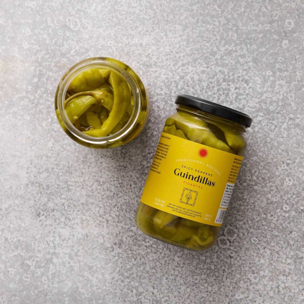 To glas med Grøndals Spicy guindillas peber, en klassisk spansk snackpeber, der er ideel til tapas, står på en lysegrå, struktureret overflade. Det ene glas er åbent, det andet har en gul produktetiket.
