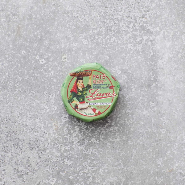 En lille, rund dåse Lucas Spicy makrelpâte med grøn emballage, rød og hvid skrift og en illustration af en person i traditionelt tøj hviler på en tekstureret grå overflade. Med et strejf af krydret tomat for ekstra smag.