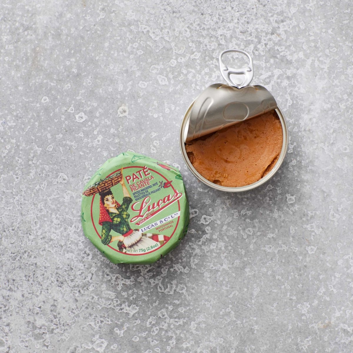 En åbnet dåse Lucas Spicy makrelpaté ligger på en grå, tekstureret overflade ved siden af det grønne låg, som har en illustration af en mand i traditionelt tøj, der holder et fad.