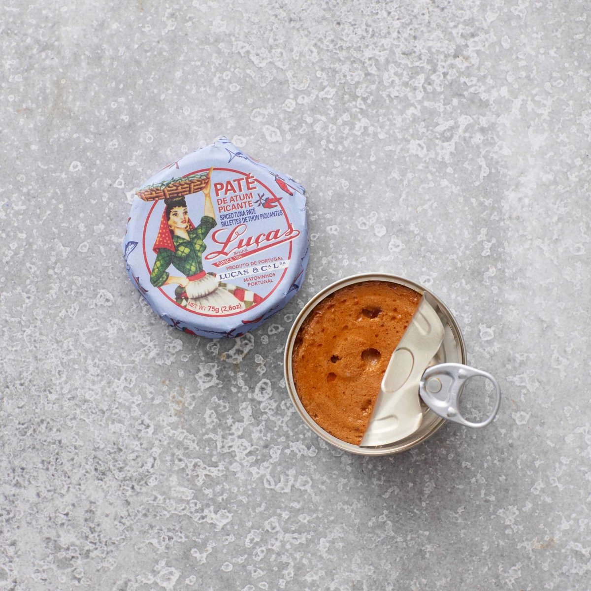 En åbnet dåse Lucas Spicy tunpaté hviler på en tekstureret grå overflade, med det illustrerede låg i vintage-stil, der viser brandingen placeret ved siden af dåsen.