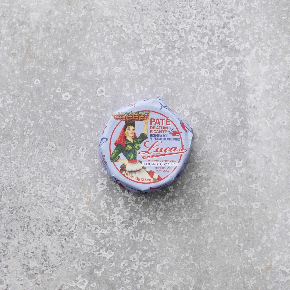 En lille rund dåse Lucas Spicy tunpaté med en vintage-etiket, der viser en kvinde, der bærer en kurv på hovedet, placeret på en tekstureret grå overflade.