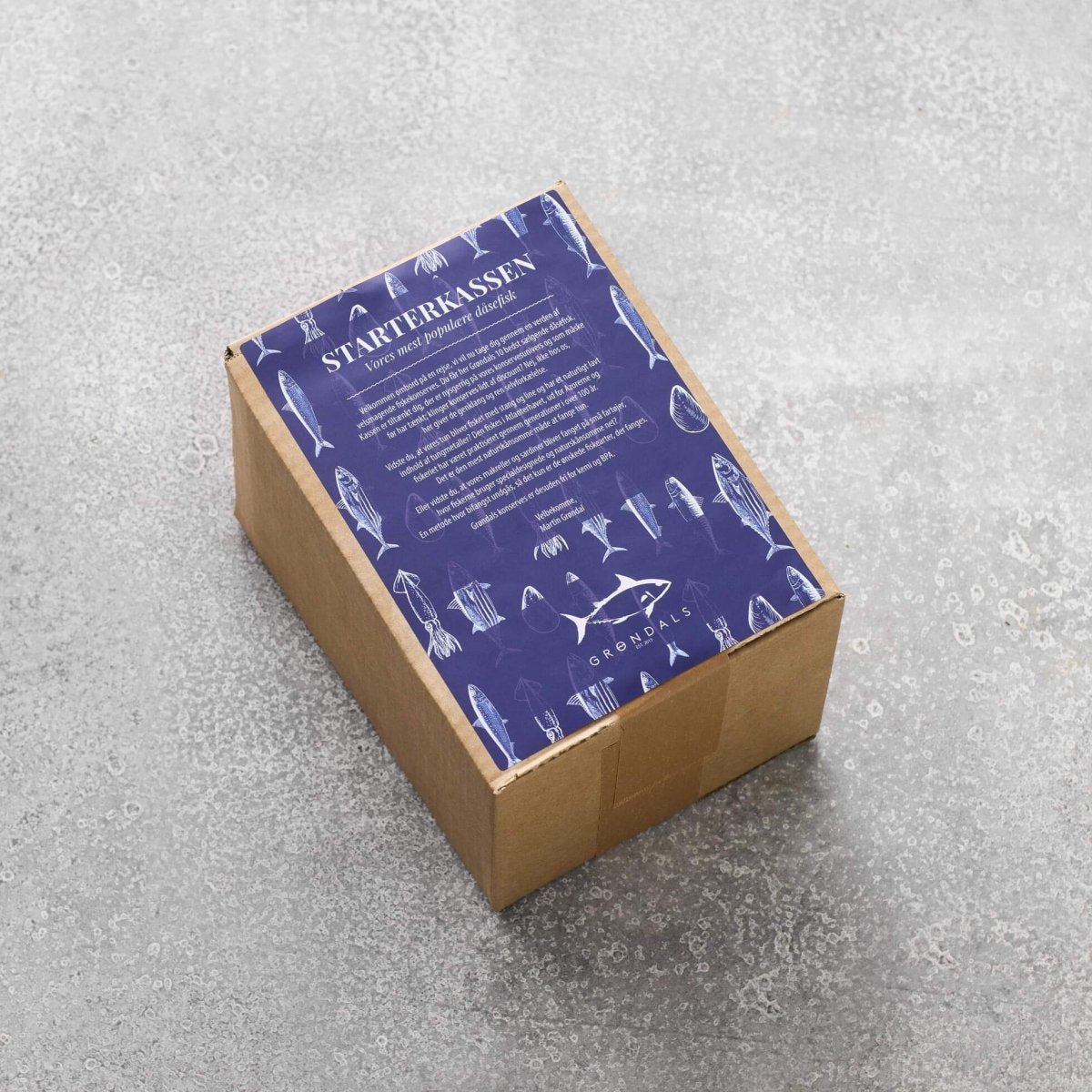 En papkasse mærket "Starterkassen" fra Grøndals med en blå top med hvid tekst og fiskeillustrationer, der fremhæver dåsefisk af høj kvalitet og yderligere produktoplysninger med mindre skrift.