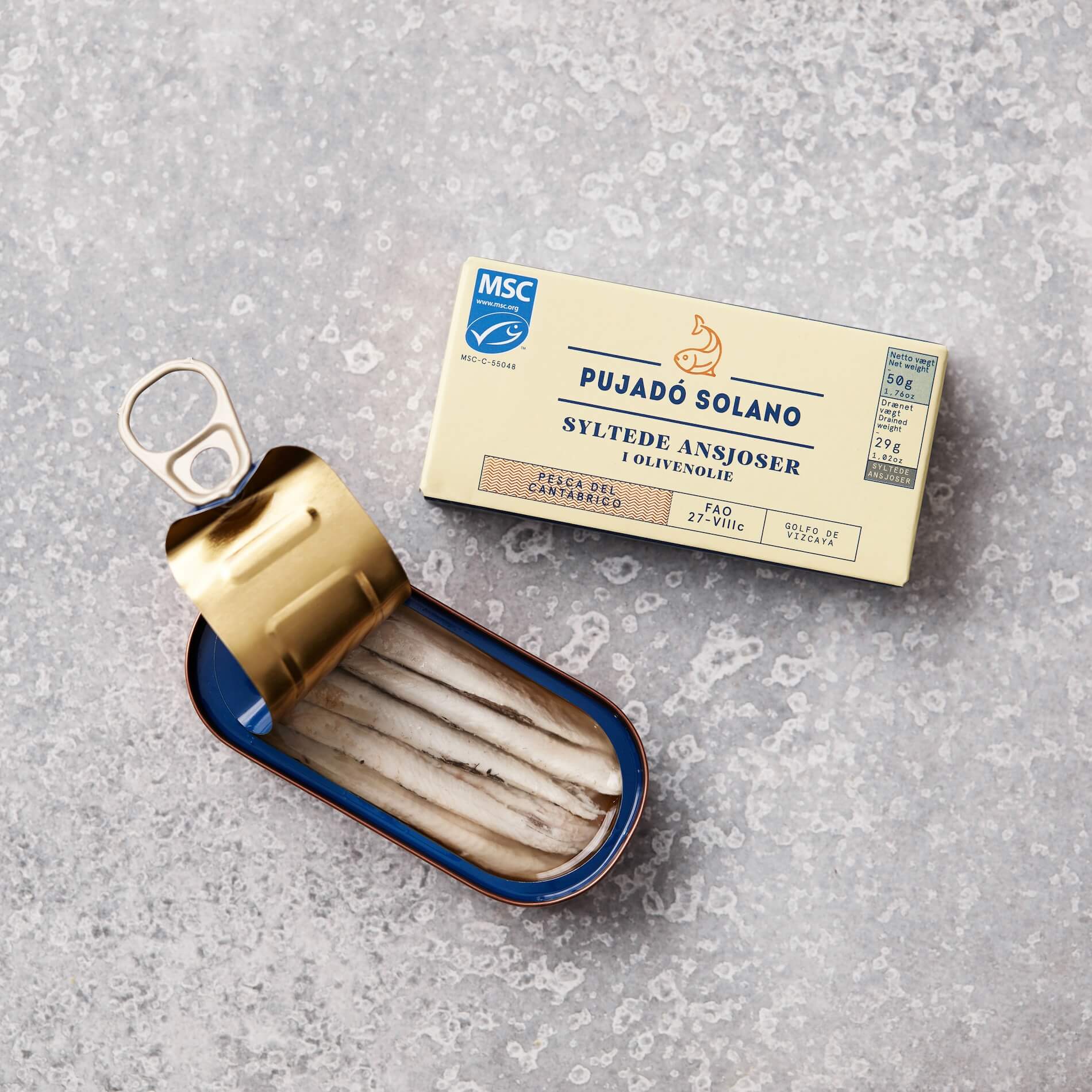En åben dåse Pujadó Solano syltede ansjoser i olivenolie fra Grøndals, præsenteret sammen med produktets emballage på en lys grå baggrund. Dåsen viser ansjosfileterne pænt arrangeret i olivenolie. Produktet er MSC-certificeret og kommer fra Cantabrico-regionen.