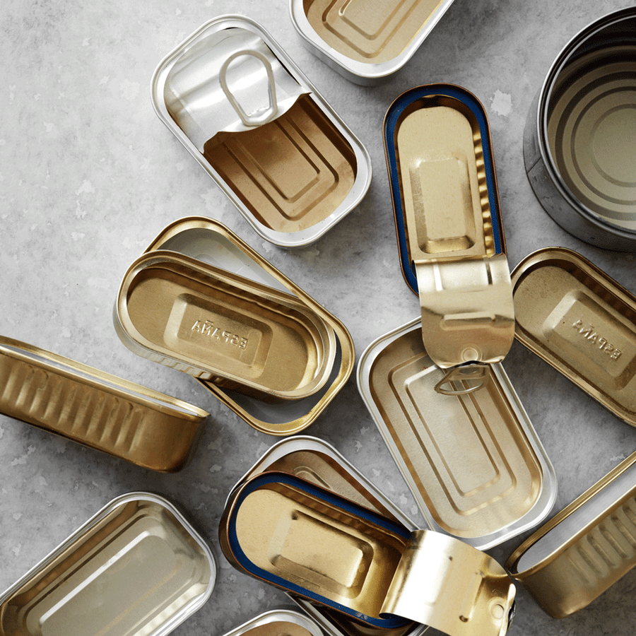 En samling åbnede, tomme metaldåser, herunder forskellige størrelser af rektangulære og runde dåser, er spredt på en lysegrå overflade. Lågene på dåserne er trukket tilbage og afslører et rent indre.