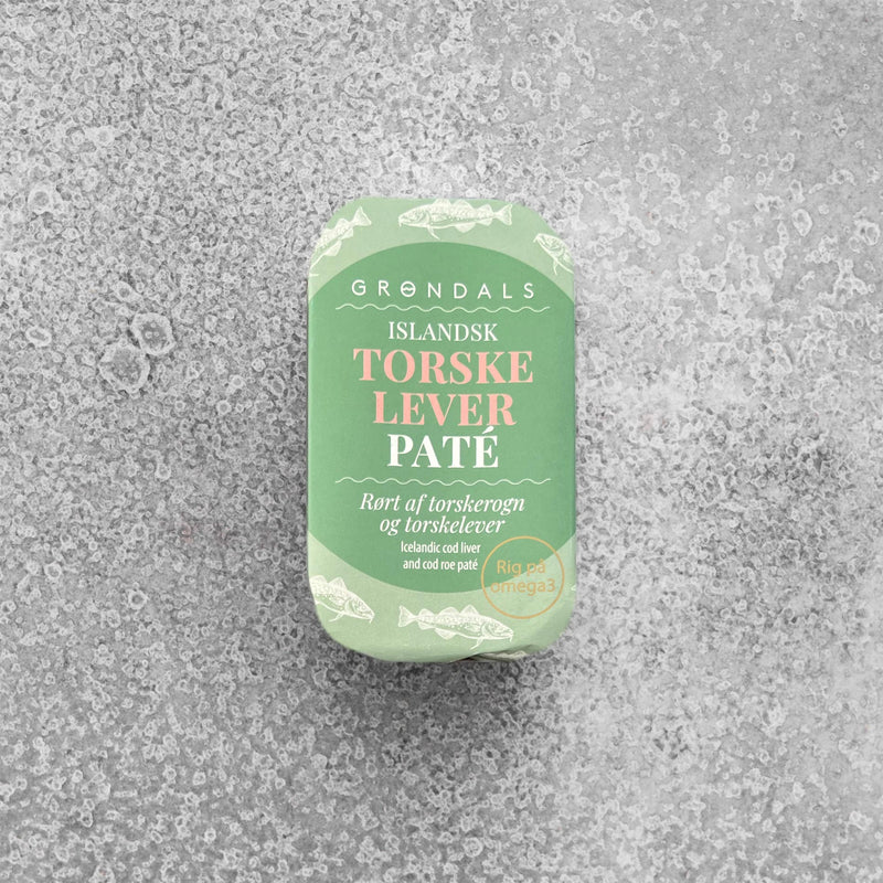 En grøn rektangulær dåse med teksten “GRØNDALS ISLANDSK TORSKELEVER PATÉ” ligger på en lysegrå, tekstureret overflade. Emballagen fremhæver indholdet som rørt af torskerogn og torskelever, rig på omega-3, og dekoreret med fiskegrafik og dansk-engelsk tekst.