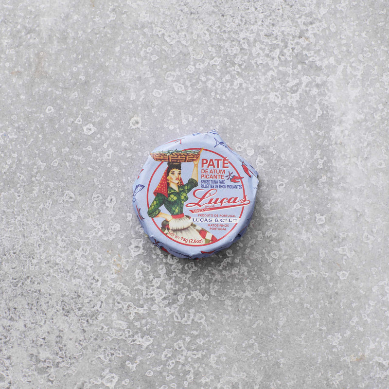 En lille rund dåse Lucas Spicy tunpaté med en vintage-etiket, der viser en kvinde, der bærer en kurv på hovedet, placeret på en tekstureret grå overflade.