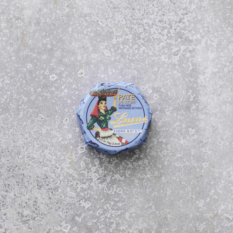 En lille, rund dåse Lucas Tunpaté med olivenolie med blå indpakning, der viser en person, der bærer en kurv på hovedet, placeret på en tekstureret grå overflade.