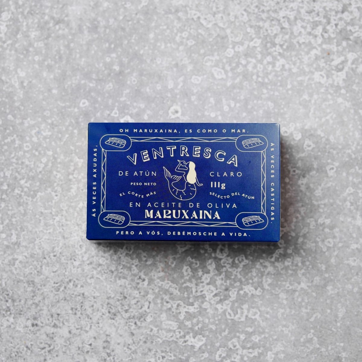En blå rektangulær dåse med Don Gastronom Tun ventresca i olivenolie hviler på en spættet grå overflade med hvid tekst og en illustration af en kvinde med fisk på emballagen.