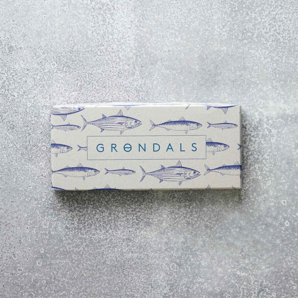 En rektangulær æske mærket "Æske til Værtsgave" fra Grøndals Praktik med et fiskemønster og "GRØNDALS" i blåt, placeret på en struktureret grå overflade.