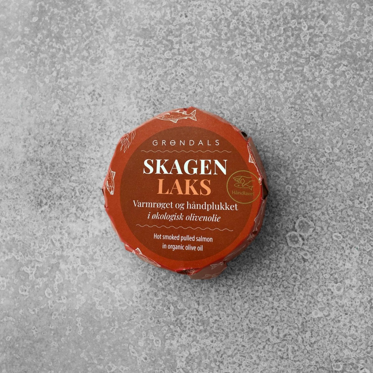 En rund, orange pakke med etiketten Grøndals Varmrøget håndplukket laks i økologisk olivenolie ligger på en grå, struktureret overflade og indikerer, at den indeholder varmrøget, håndplukket laks i økologisk olivenolie.