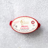 Oval dåse med Pulpo i galisisk sauce fra Conservas De Cambados med en fløde- og rød etiket med gourmetbranding. Dåsen indeholder mørt blækspruttearme i fyldig galisisk sauce, præsenteret på en tekstureret grå overflade med røde accenter.