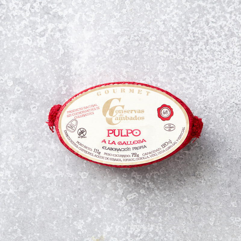 Oval dåse med Pulpo i galisisk sauce fra Conservas De Cambados med en fløde- og rød etiket med gourmetbranding. Dåsen indeholder mørt blækspruttearme i fyldig galisisk sauce, præsenteret på en tekstureret grå overflade med røde accenter.