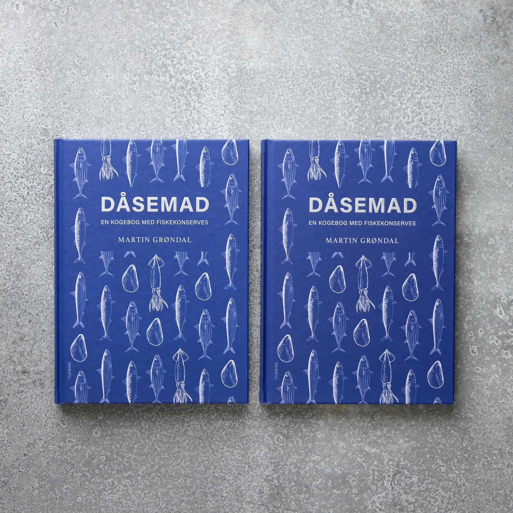 To blå indbundne eksemplarer af Grøndals' kogebog "DÅSEMAD" ligger side om side på en grå tekstureret overflade. Omslaget har hvide konturer af fisk og skaldyr, der antyder opskrifterne på konserves til fisk og skaldyr indeni.