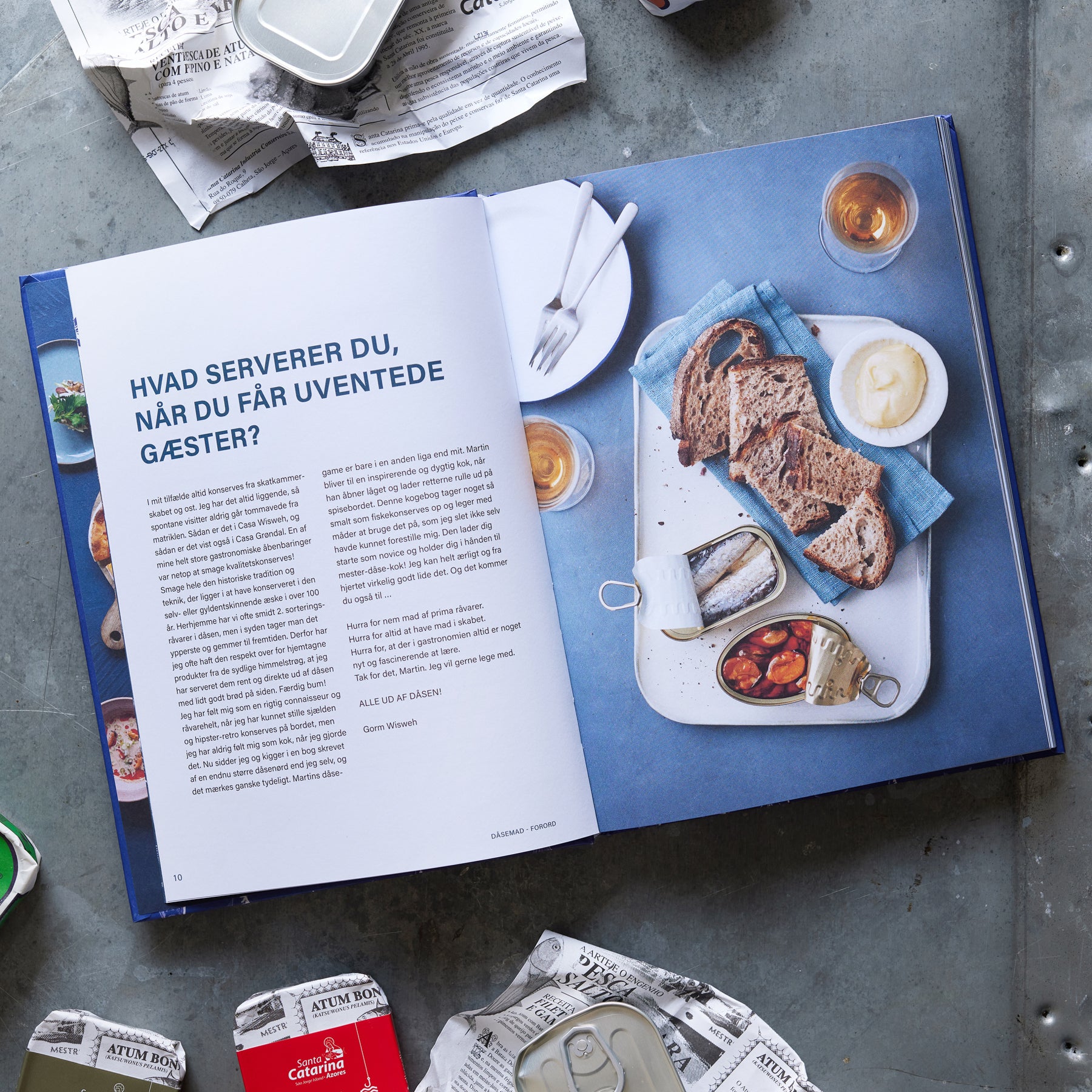 Grøndals' kogebog, Kogebogen DÅSEMAD, står åben med danske opskrifter ved siden af en hvid bakke med rugbrød, smør, fiskekonfitører og drikkevarer, omgivet af avispapir og tomme dåser.