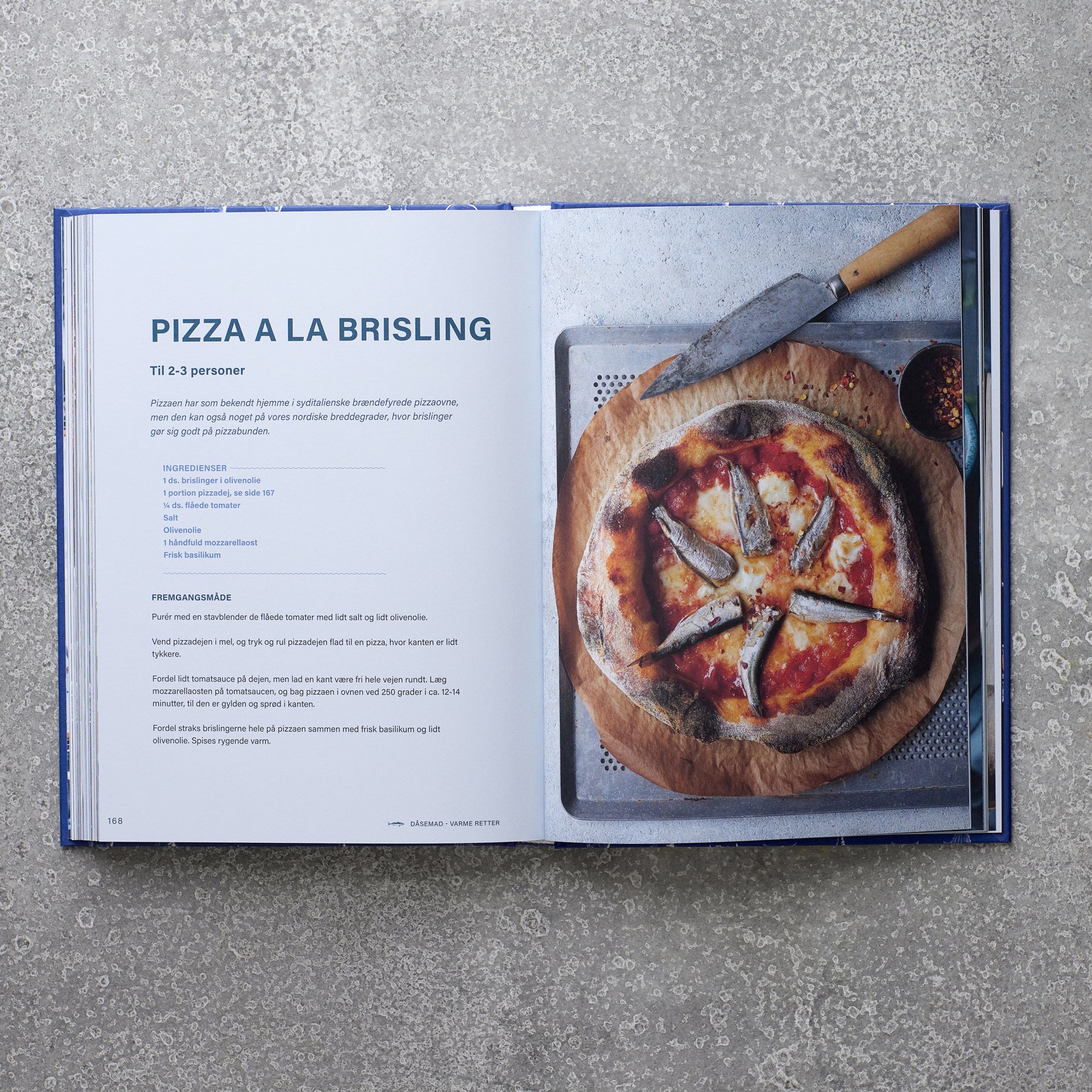 Et åbent eksemplar af Grøndals' Kogebogen DÅSEMAD viser opskriften på Pizza a la Brisling til venstre, mens højre side viser en bagt pizza med fiskekonserves, placeret på en bakke med en kniv ved siden af.