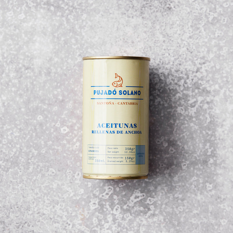 En dåse Fyldte oliven med ansjoser står på en lysegrå, spættet overflade. Den cremefarvede etiket med blå og gylden tekst fremhæver umamismagen og de eksklusive kvaliteter ved disse fyldte oliven.
