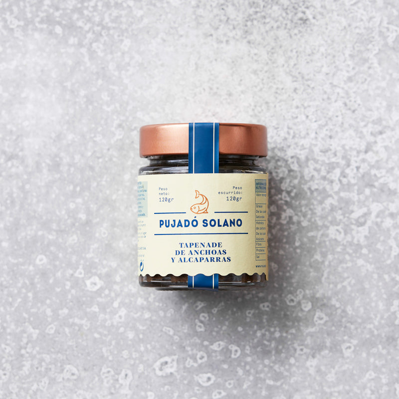 Et glas Pujado Solano Tapenade af ansjos, oliven og kapers står på en lysegrå tekstureret overflade med et kobberfarvet låg – en smagfuld tilføjelse fra middelhavskøkkenet.