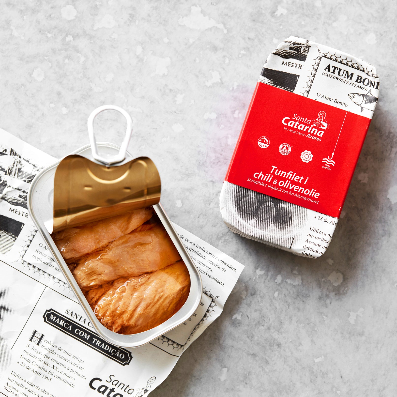 En åbnet dåse tunfilet ligger på trykt papir ved siden af sin rød-hvide emballage med teksten “Tunfilet i mild chili og olivenolie”, med et strejf af azorisk chili, alt sammen anrettet på en lysegrå overflade.
