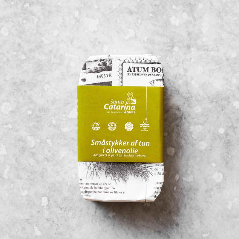 En rektangulær dåse Santa Catarina Småstykker af tun i økologisk olivenolie, pakket ind i avispapir med en grøn dansk etiket, er placeret på en lysegrå tekstureret overflade.
