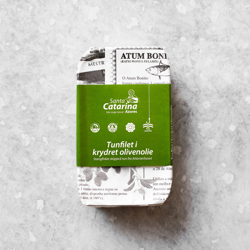 En rektangulær dåse Tunfilet i krydret olivenolie, indpakket i grøn- og hvidt papir, ligger på en lysegrå, tekstureret overflade.
