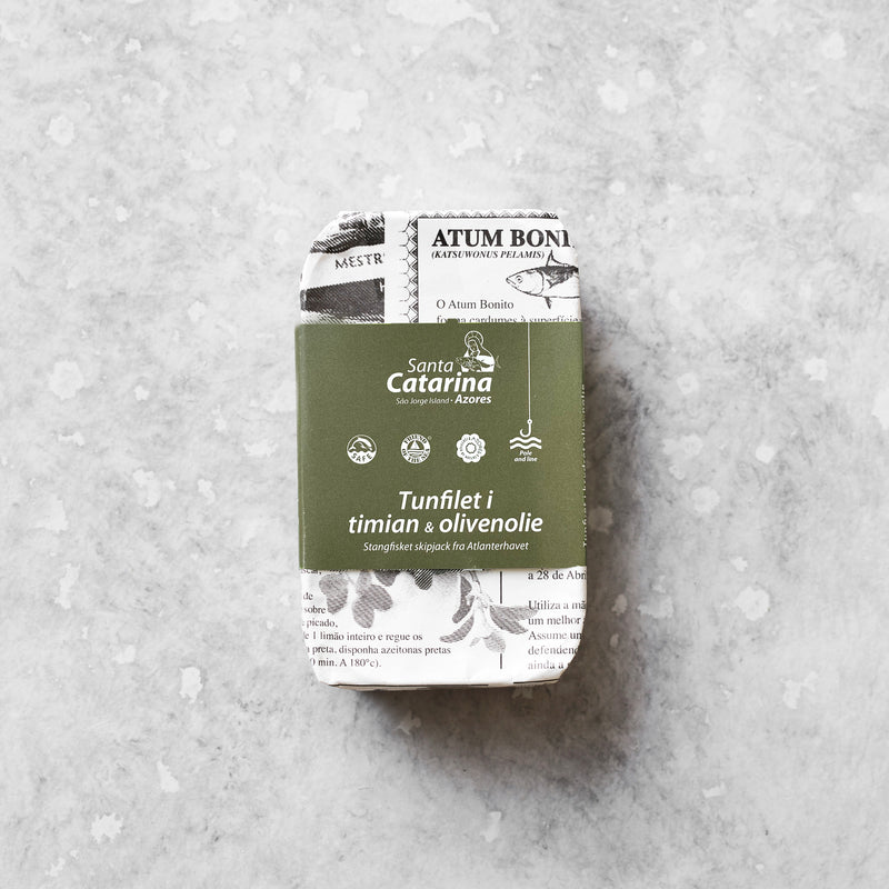 En dåse Santa Catarina Tunfilet i timian og olivenolie, pakket ind i grønt og hvidt papir med vintagetekst og illustrationer, hviler på en lysegrå stenoverflade.
