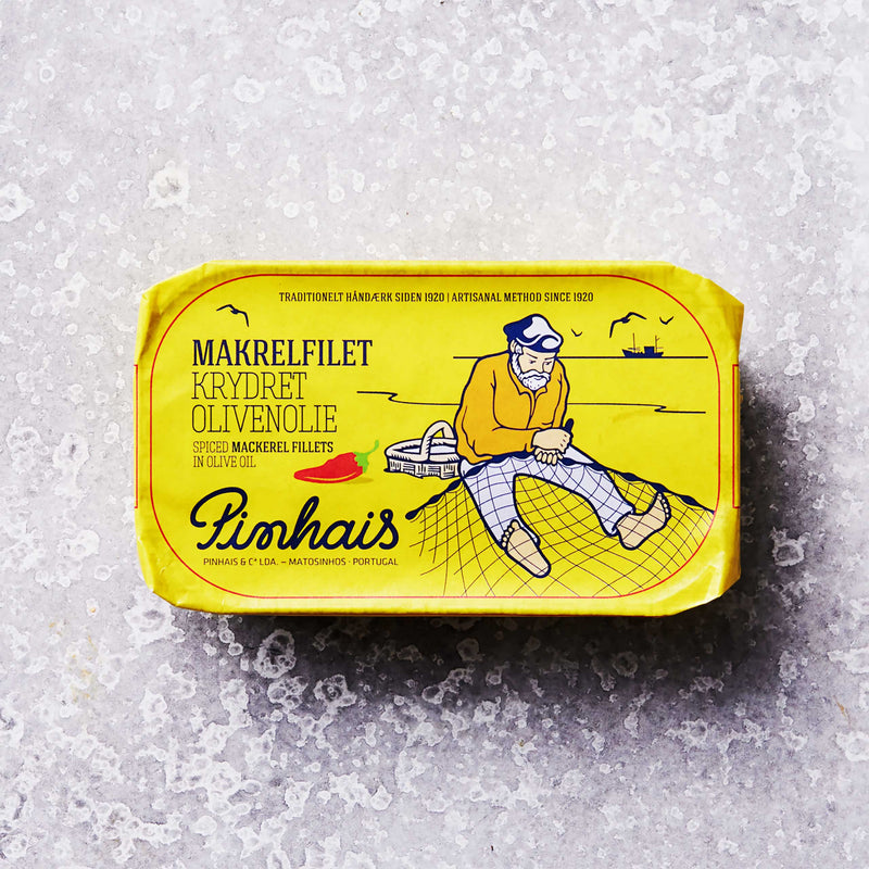 En gul dåse Pinhais Makrelfilet i krydret olivenolie med illustration af en fisker på et net og en båd. Dansk og engelsk tekst fremhæver bæredygtigt fiskeri.