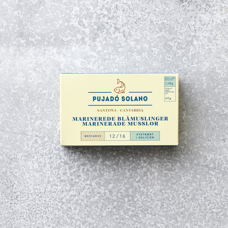 En cremefarvet rektangulær æske med Pujado Solano Muslinger i escabeche, en middelhavsdelikatess, med blå og guld accenter hviler på en lysegrå tekstureret overflade.