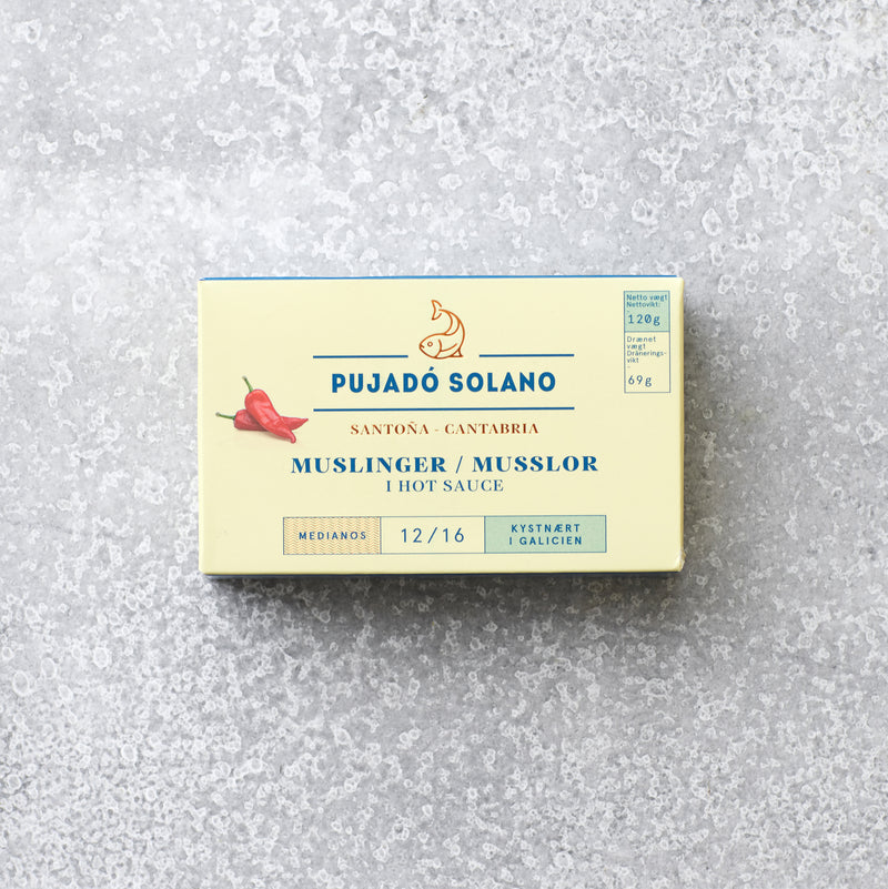 En rektangulær dåse Pujado Solano muslinger i hot sauce, dekoreret med to røde chilipeber og blågylden tekst på en lys baggrund, ideel til dit tapasbord og vist på en tekstureret grå overflade.