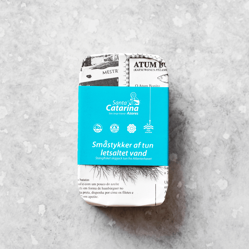 En dåse Santa Catarina Tun i letsaltet vand, pakket ind i sort-hvidt papir med en blå etiket med dansk tekst og ikoner, er placeret på en lysegrå, spættet overflade.