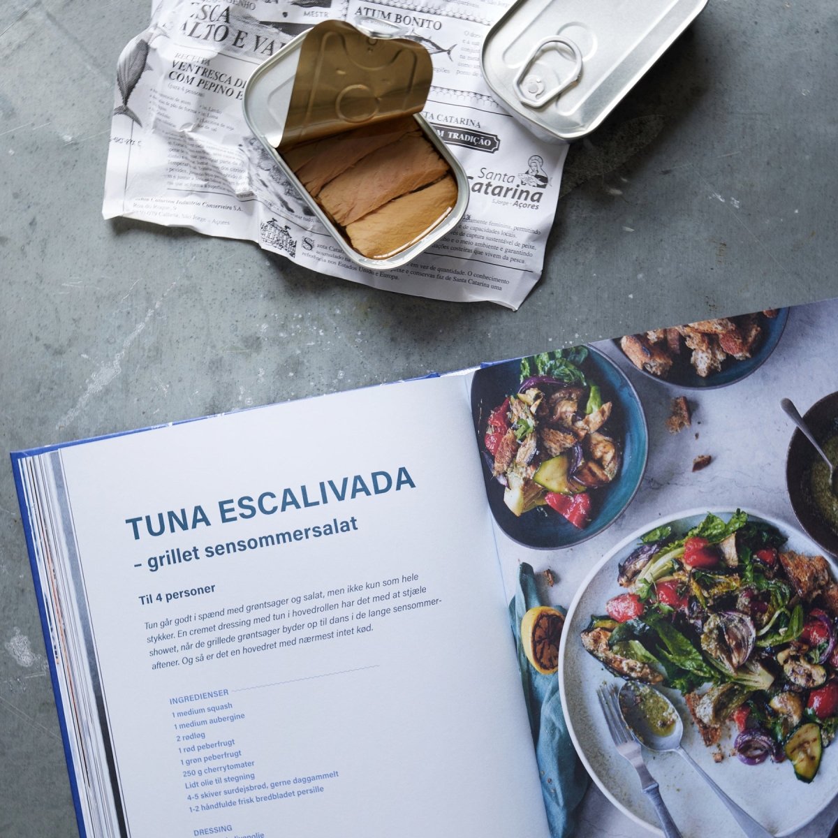 En åben kogebog med en opskrift på Tuna Escalivada ligger ved siden af en salat og en åbnet dåse fra Gavekasse og DÅSEMAD fiskekonserves på avispapir, alt sammen anrettet på en grå bordplade.