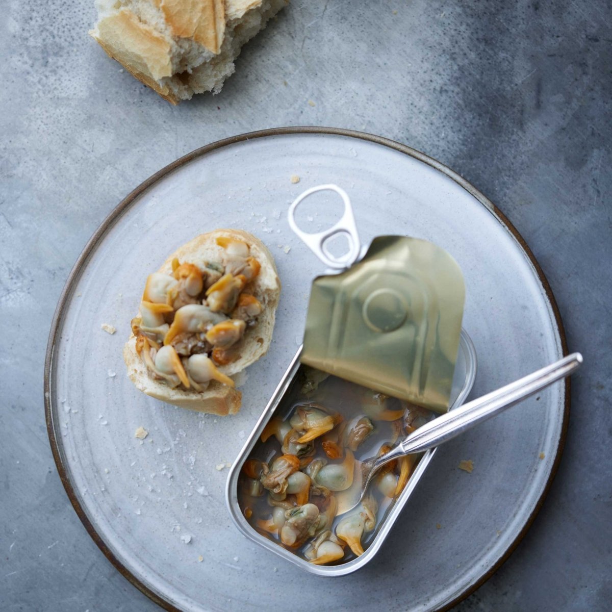 En tallerken holder en skive brød toppet med "Don Gastronom Hjertemuslinger i egen lage" ved siden af en åben dåse med muslinger i lage og en gaffel. Et stykke brød hviler på tallerkenens kant, helt nede på en grå overflade. Bæredygtig Galicien skaldyr.