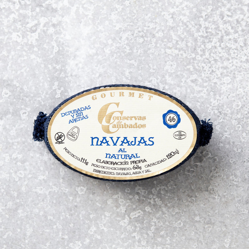 En dåse Grøndals Knivmuslinger i letsaltet lage hviler på en lysegrå tekstureret overflade. Den ovale etiket viser blå og gylden spansk tekst med ingredienser og produktoplysninger.