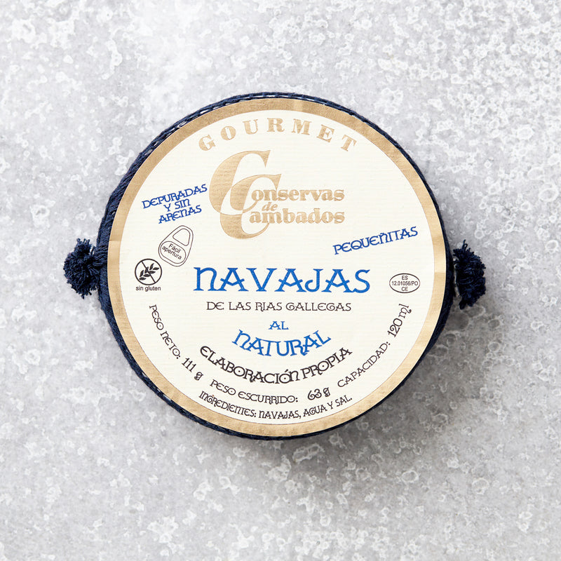 Rund dåse med Små knivmuslinger i letsaltet lage fra Conservas De Cambados med blå og guld tekst på et trætekstureret låg, sat på en lys stenbaggrund.