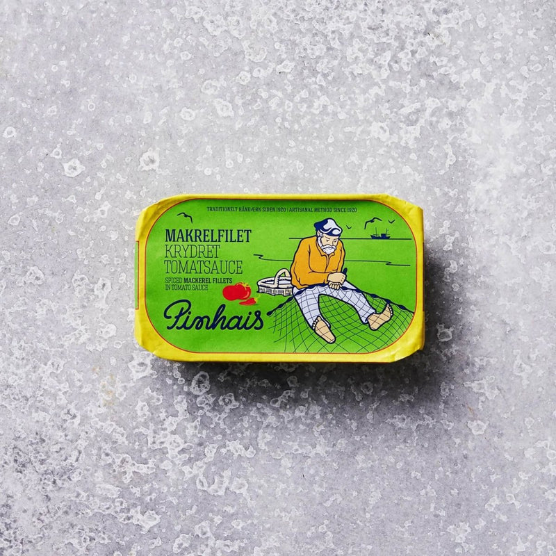 En dåse Pinhais Makrelfilet i krydret tomatsauce med en grøn etiket, der viser en skægget mand i gult på et fiskenet og en båd i baggrunden.