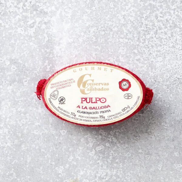 Oval dåse med Pulpo i galisisk sauce fra Conservas De Cambados med en fløde- og rød etiket med gourmetbranding. Dåsen indeholder mørt blækspruttearme i fyldig galisisk sauce, præsenteret på en tekstureret grå overflade med røde accenter.