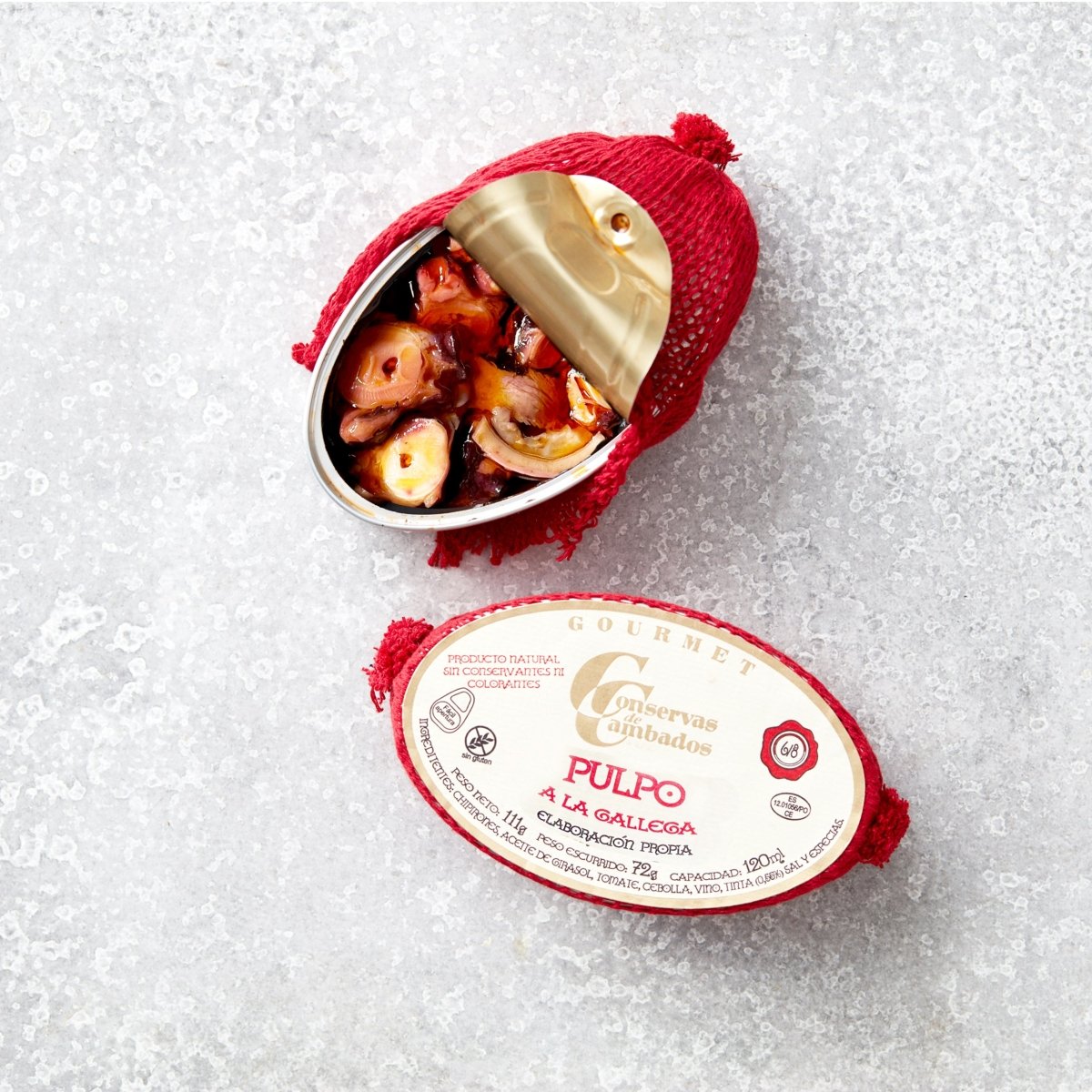 En åbnet dåse Pulpo i galisisk sauce fra Conservas De Cambados, pakket ind i rødt net, hviler på en tekstureret lysegrå overflade. Etiketten fremhæver gourmet-brandingen og produktnavnet.