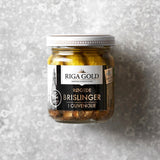 En glaskrukke med Riga Gold Røgede brislinger i olivenolie med sølvlåg og en sort-hvid etiket med tekst og en kirkeillustration hviler på en grå, struktureret overflade.