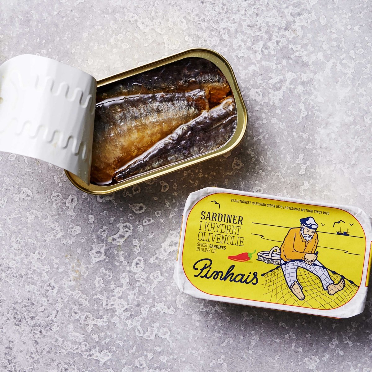 En åbnet dåse med Sardiner i krydret olivenolie fra Pinhais ligger ved siden af låget, som viser en illustration af en person på et net og etiketten "Sardiner i krydret olivenolie, Pinhais.