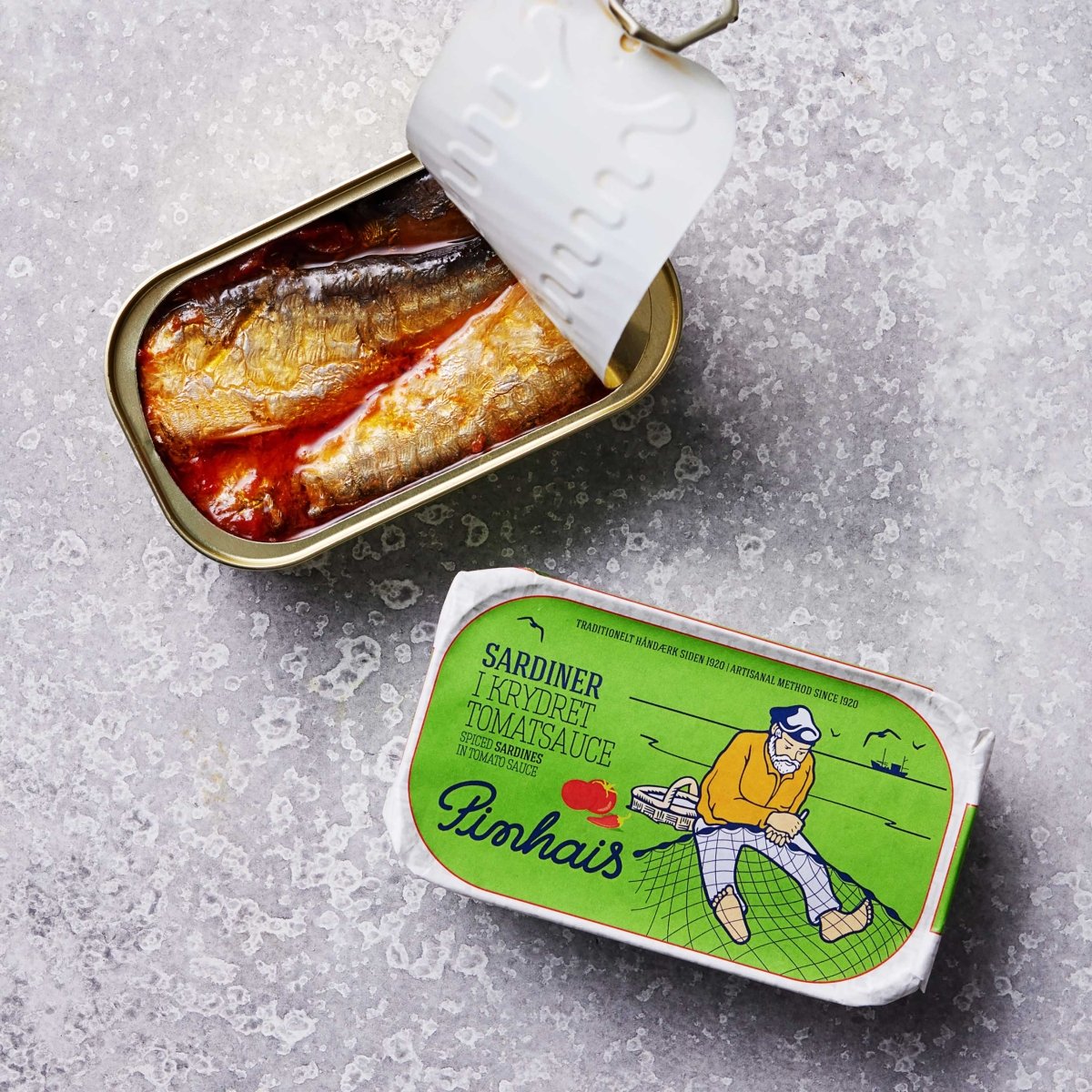 En åbnet dåse Pinhais Sardiner i krydret tomat ligger ved siden af emballagen, som viser en fiskeillustration og produktnavnet. Disse sardiner er rige på omega-3-fedtsyrer og er velsmagende og nærende.