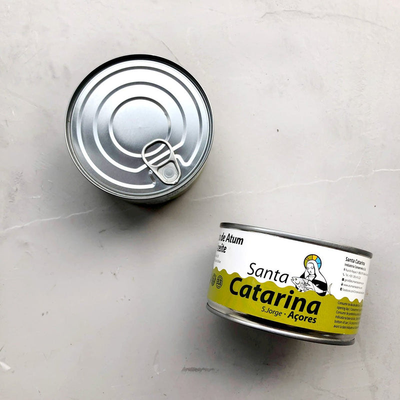 To dåser Santa Catarina Småstykker af tun i olivenolie står på en lysegrå overflade. Den ene står oprejst og viser en illustration og etiket af en nonne; den anden er på hovedet og viser sit enkle metallåg med trækflig.