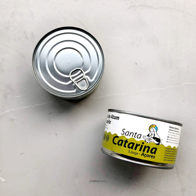 To dåser Santa Catarina Småstykker af tun i olivenolie står på en lysegrå overflade. Den ene står oprejst og viser en illustration og etiket af en nonne; den anden er på hovedet og viser sit enkle metallåg med trækflig.
