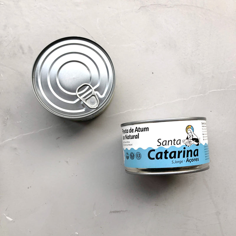 To dåser Santa Catarina Småstykker af tun i letsaltet vand hviler på en lysegrå overflade – den ene har det sølvfarvede låg med trækfane, mens den anden har den blå og hvide etiket, der fremhæver dette bæredygtige valg.