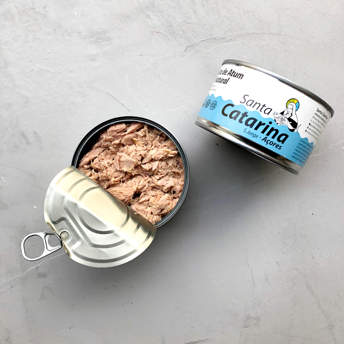 To dåser Santa Catarina Småstykker af tun i letsaltet vand; den ene forseglet, den anden delvist åbnet for at vise bæredygtige tunstykker indeni. Begge er placeret på en lysegrå overflade.