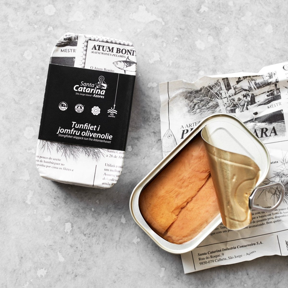 En åbnet dåse Santa Catarina Tunfilet i jomfru olivenolie hviler på vintagepapir ved siden af sin elegante sort-hvide æske, der er sat på en lysegrå sten – fanget med den bæredygtige fangstmetode.