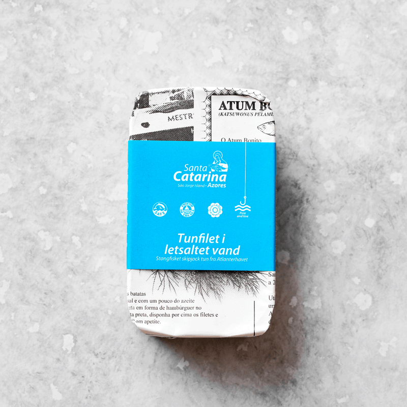En dåse Santa Catarinas Tunfilet i letsaltet vand, pakket ind i sort-hvidt papir med en blå etiket, hviler på en lysegrå, spættet overflade – et kvalitetsprodukt på dåse fra bæredygtigt fiskeri.
