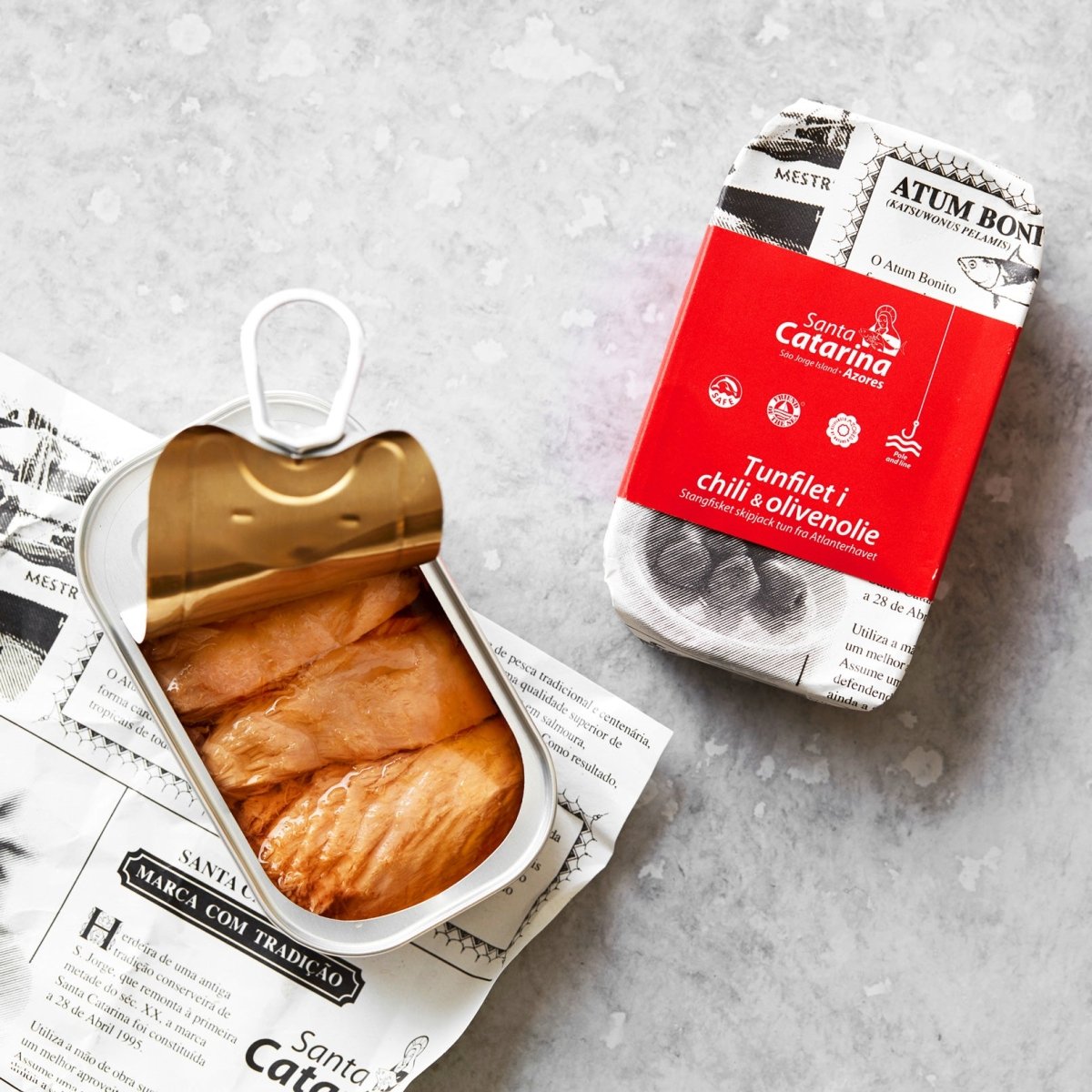 En åbnet dåse tunfilet ligger på trykt papir ved siden af sin rød-hvide emballage med teksten “Tunfilet i mild chili og olivenolie”, med et strejf af azorisk chili, alt sammen anrettet på en lysegrå overflade.
