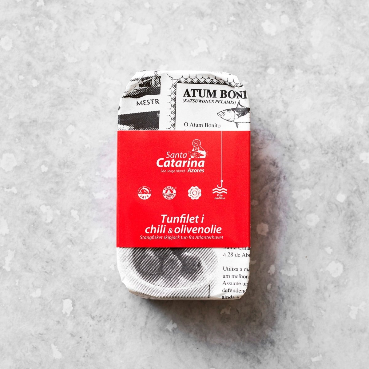 En dåse Tunfilet i mild chili og olivenolie, indpakket i papir med sort-hvid tekst og en rød etiket, ligger på en lys, tekstureret baggrund.
