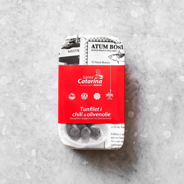 En dåse Tunfilet i mild chili og olivenolie, indpakket i papir med sort-hvid tekst og en rød etiket, ligger på en lys, tekstureret baggrund.
