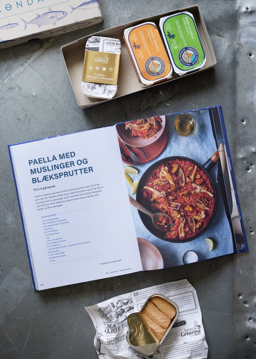 En åben kogebog med paella står på et metalbord sammen med Grøndals Værtsgave og DÅSEMAD, inklusive muslinger og fiskedåser. I nærheden er et billede ovenfra af paella i en pande og åbne dåser på avispapir.