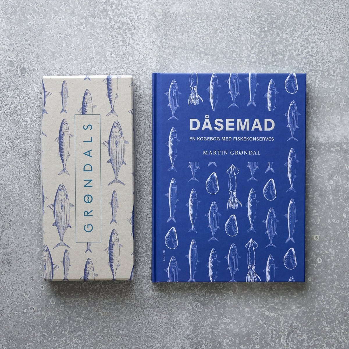 En blå DÅSEMAD kogebog med fiskeillustrationer og en beige Grøndals Værtsgave-æske, begge dekoreret med fiskekonserves-tegninger, er placeret på en spættet grå overflade.